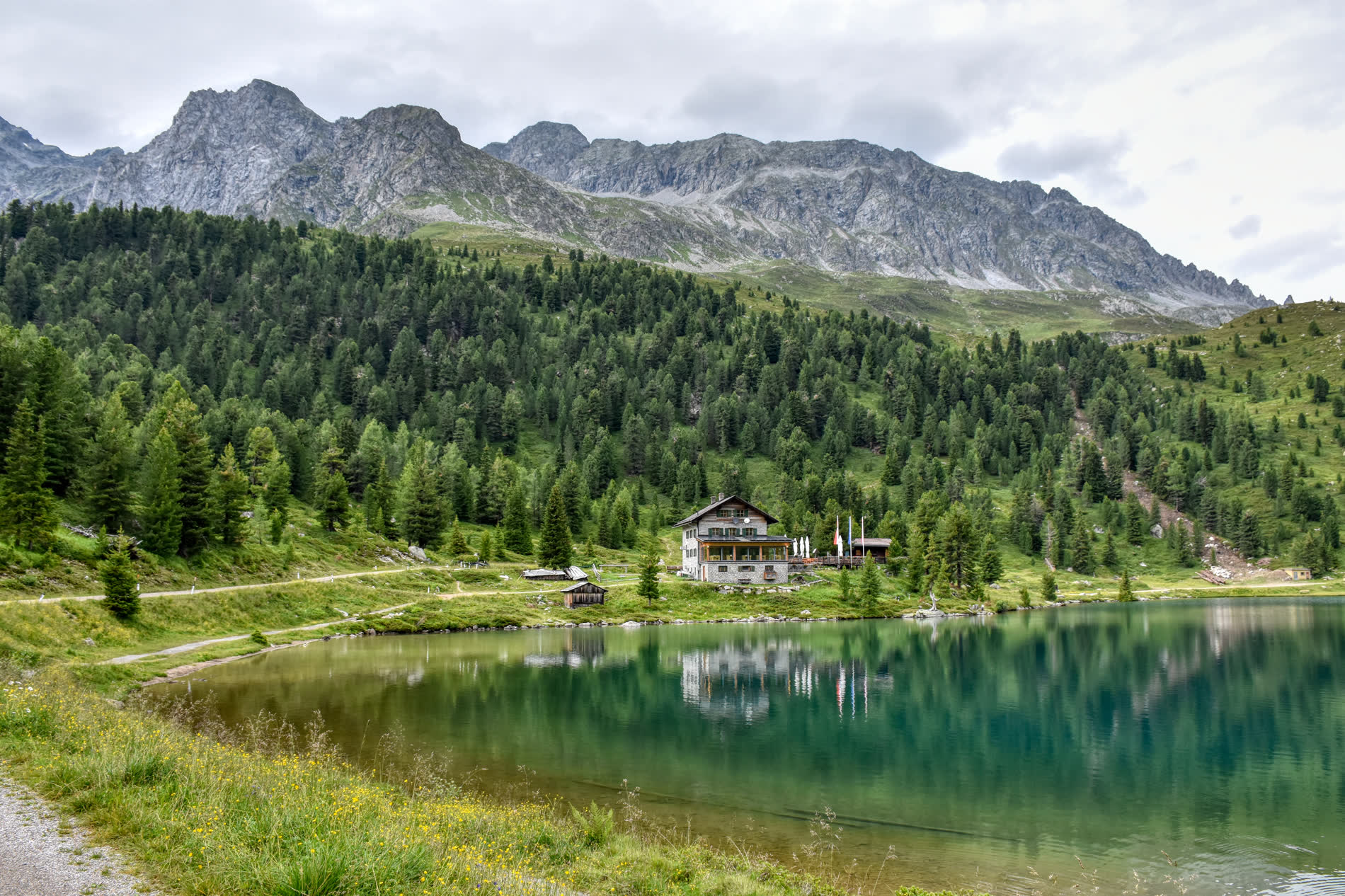 Grüner Bergsee vor Fichtenwald, am Ufer ein Gasthaus, dahinter ein felsiger Grat Grüner Bergsee vor Fichtenwald, am Ufer ein Gasthaus, dahinter ein felsiger Grat